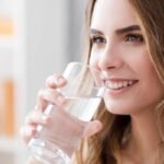 A smiling woman in a modern kitchen drinks a glass of water, demonstrating the benefits of daily hydration and a healthy lifestyle